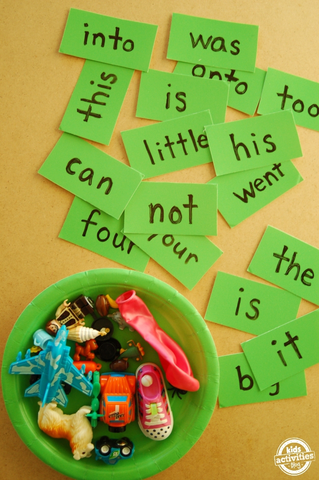 Sight words written on green cards on and spread out on a yellow table next to a green bowl of various toys.